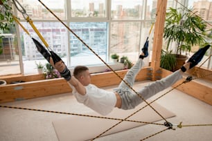 Junger Sportler in Aktivkleidung über Matte auf dem Boden hängend, seine Arme und Beine durch spezielle Ausrüstung mit Seilen fixiert
