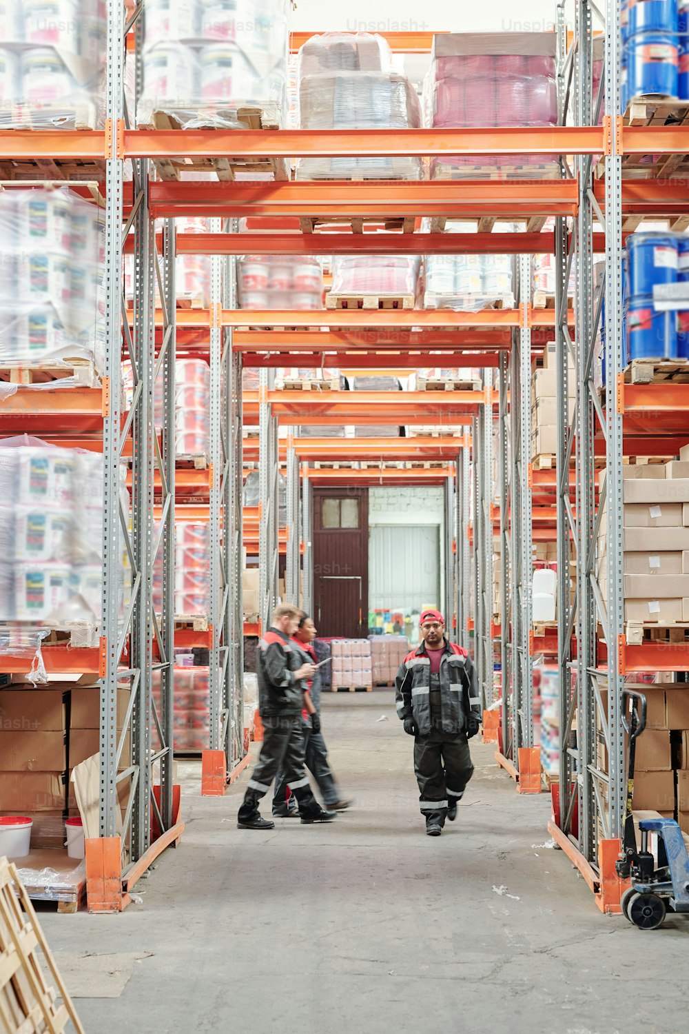 Young mixed-race male warehouse worker in uniform moving along aisle between large racks while his colleagues walking behind him
