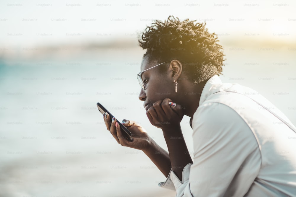 Seitenansicht einer entzückenden schwarzen Frau in Brille und weißem Trench, mit lockigem Afro-Haar, Ohrringen und Nagelkunst liest an einem sonnigen Tag nachdenklich einen Feed sozialer Netzwerke im Freien