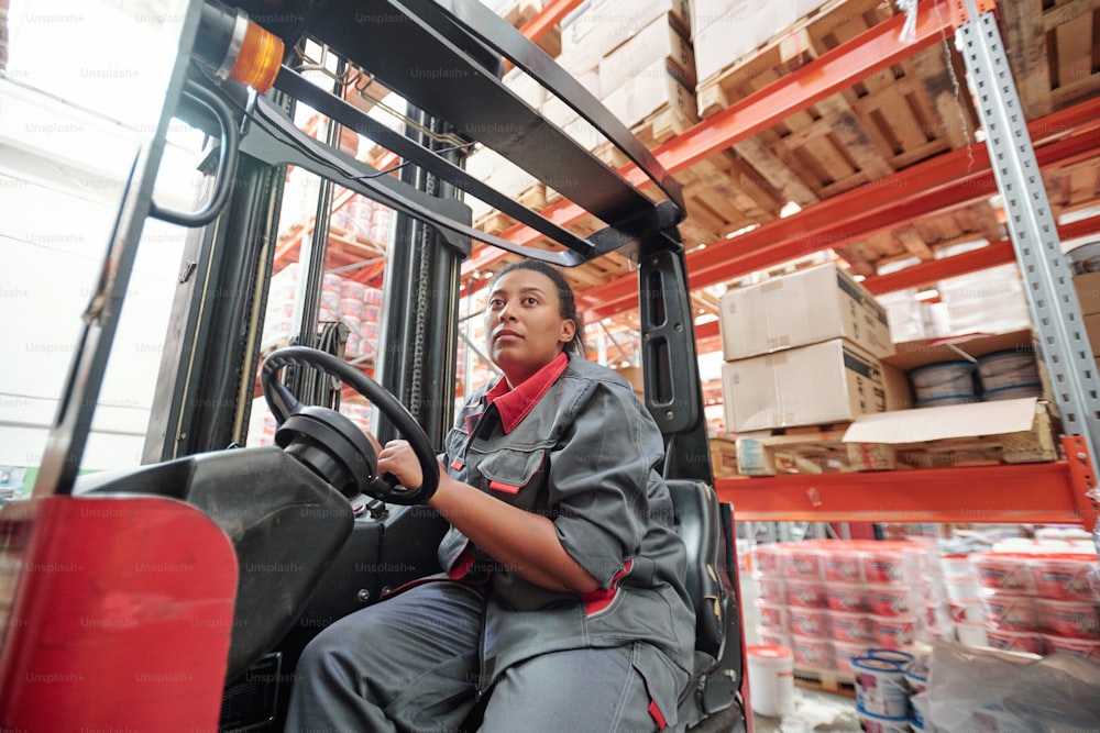 Young mixed-race woman in workwear sitting by steer inside industrial machine during work in warehouse on background of racks