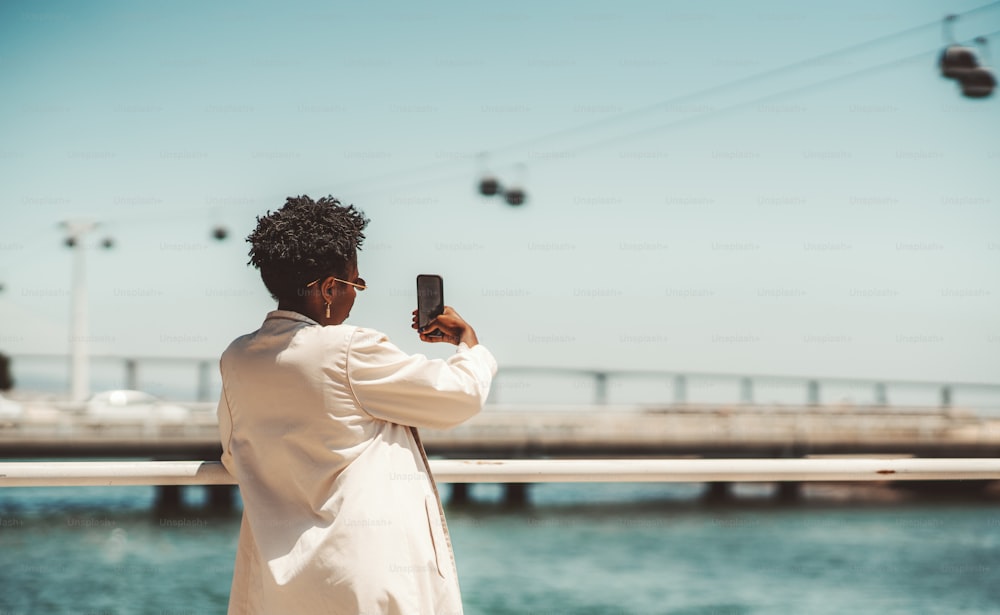 View from behind of a young black female tourist with a curly Afro hair, in a white trench and curly afro hair taking pics of sightseeing and ropeway over the water; a copy space place on the right