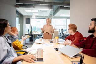 Femme blonde mature en tenue décontractée debout près de la table devant ses jeunes collègues interculturels lors d’une formation commerciale au bureau