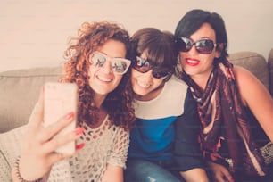 Three female friends at home use modern cellular phone to take selfie or do video call with people - cheerful trendy young women in indoor communication activity