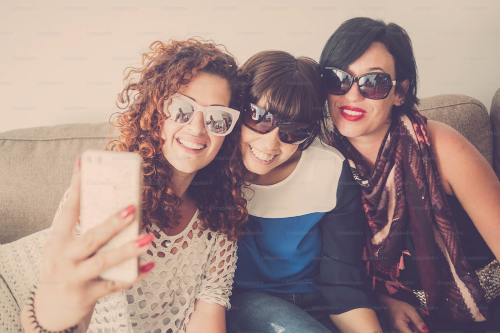 Three female friends at home use modern cellular phone to take selfie or do video call with people - cheerful trendy young women in indoor communication activity