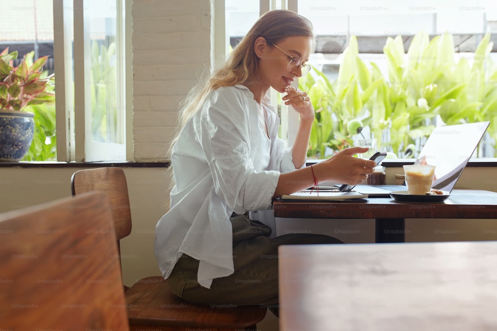 Work. Woman At Cafe Using Smartphone. Happy Stylish Girl In Glasses Reading And Touching Chin. Laptop And Digital Technologies For Remote Working Or Online Studying At Comfortable Workplace.