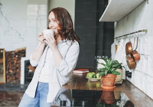 Adult smiling brunette woman in casual with cup of tea on kitchen at home