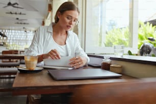 Business. Woman At Cafe Working Remotely. Girl In Casual Clothes And Glasses With Laptop Writing Something In Notebook. Digital Technologies For Creative Job At Comfortable Place On Summer Vacation.