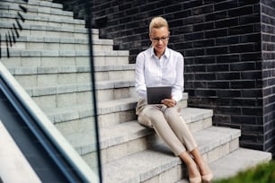 Attractive blond smiling fashionable businesswoman sitting on the stairs outdoors and using tablet.