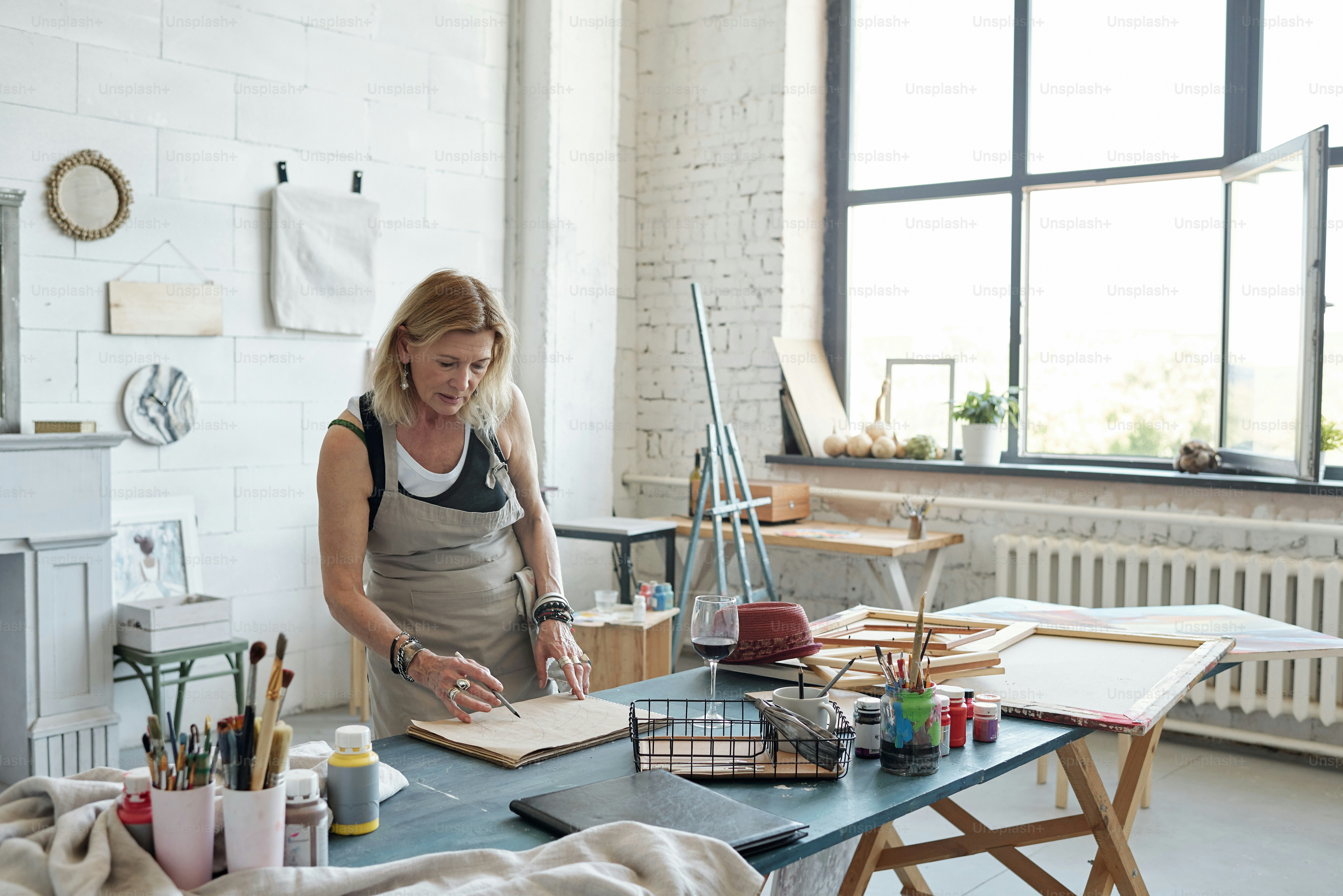 Attractive mature woman in apron standing at table and working on sketch in art studio