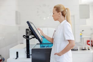 Hermosa asistente de laboratorio rubia de pie en el laboratorio y usando la computadora para ingresar los resultados de las pruebas.