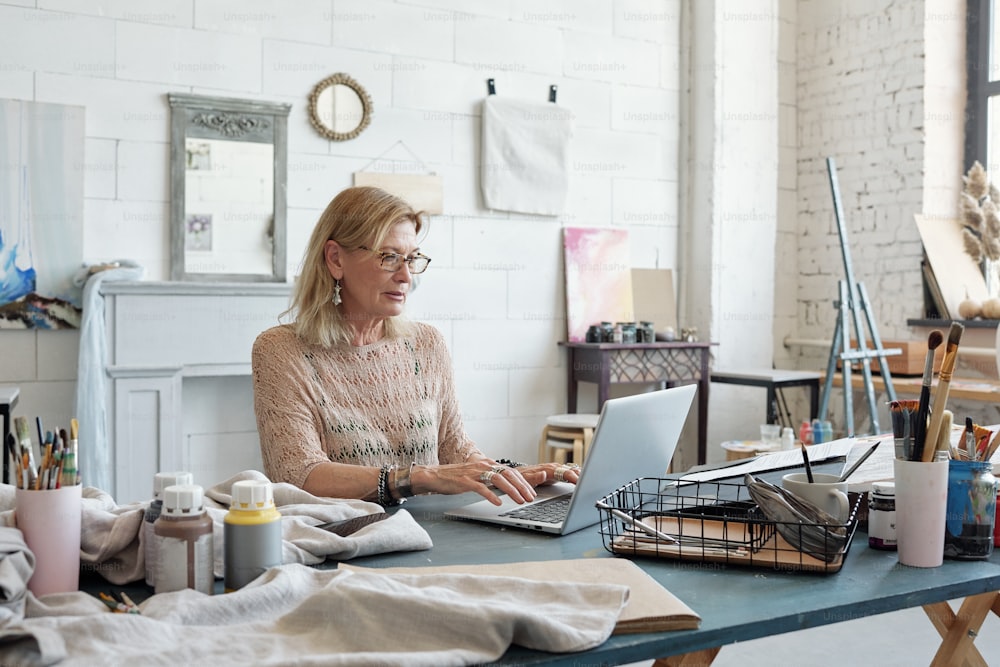 금발 머리를 가진 바쁜 여성 아티스트가 테이블에 앉아 자신의 스튜디오에서 노트북에 타이핑하고 있습니다.