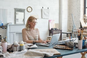 Busy female artist with blond hair sitting at table and typing on laptop in own studio