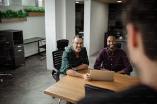 Due uomini d'affari diversi sorridenti che parlano con un collega mentre lavorano insieme a un tavolo in un ufficio