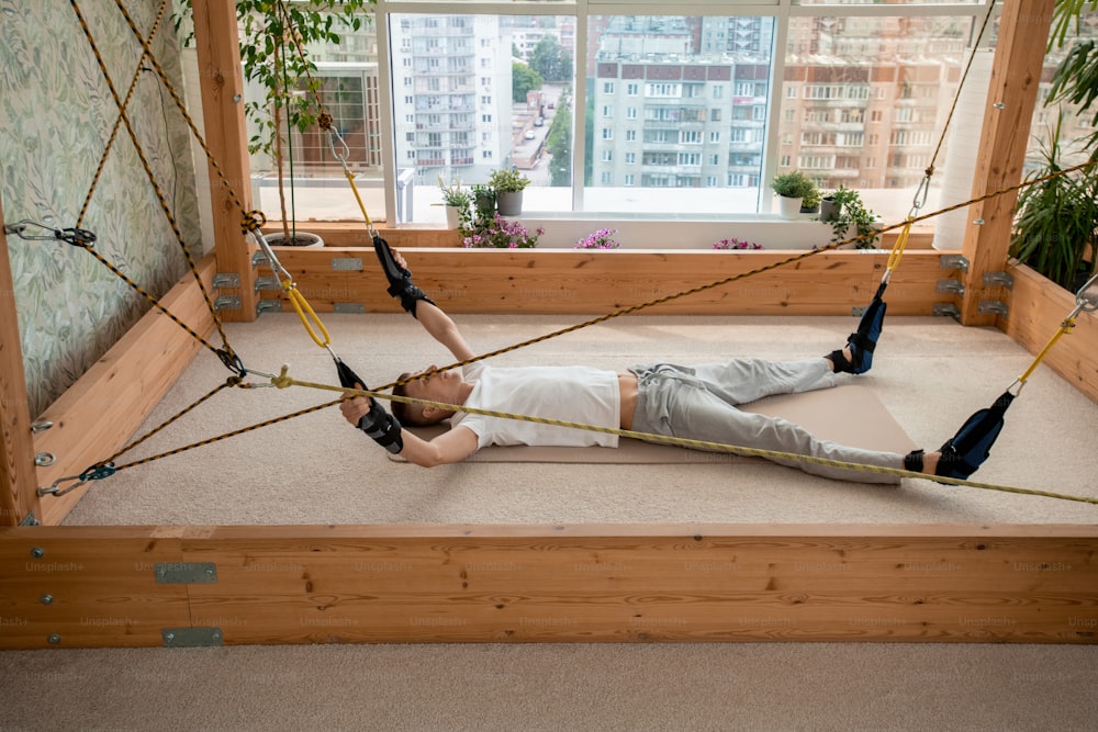 Young sportsman in activewear lying on mat inside rectangular wooden structure with his arms and legs fixed by special equipment and ropes