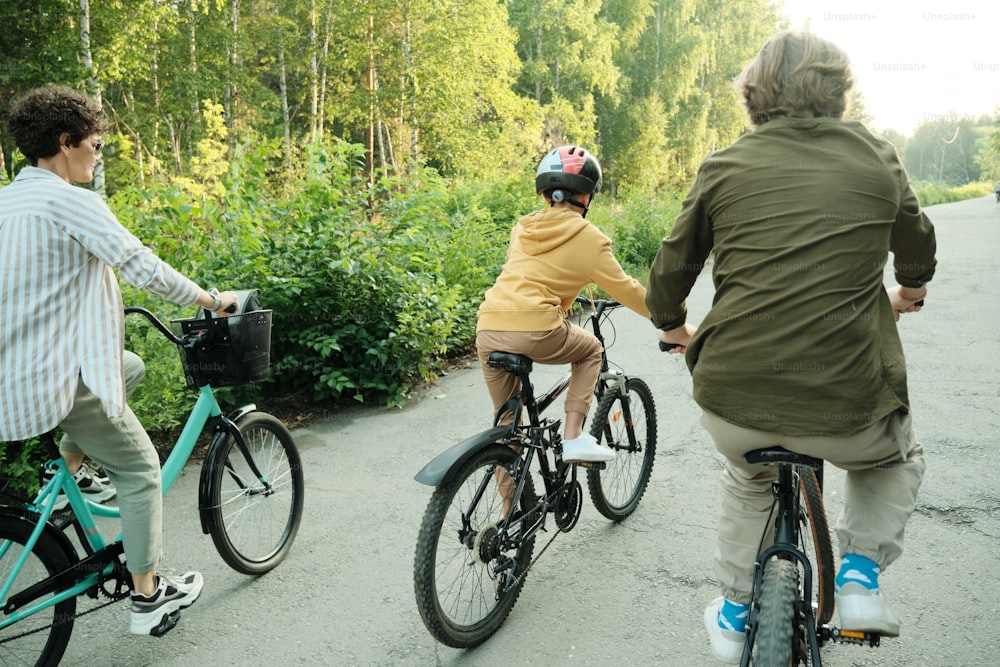 Famiglia attiva di marito, moglie e il loro figlio di età elementare che vanno in bicicletta lungo la strada nel parco moderno in una giornata di sole o mattina
