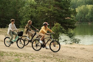 Famille contemporaine heureuse de trois personnes en tenue décontractée assise sur des vélos tout en se déplaçant le long de la rivière tout en profitant d’un repos actif ensemble