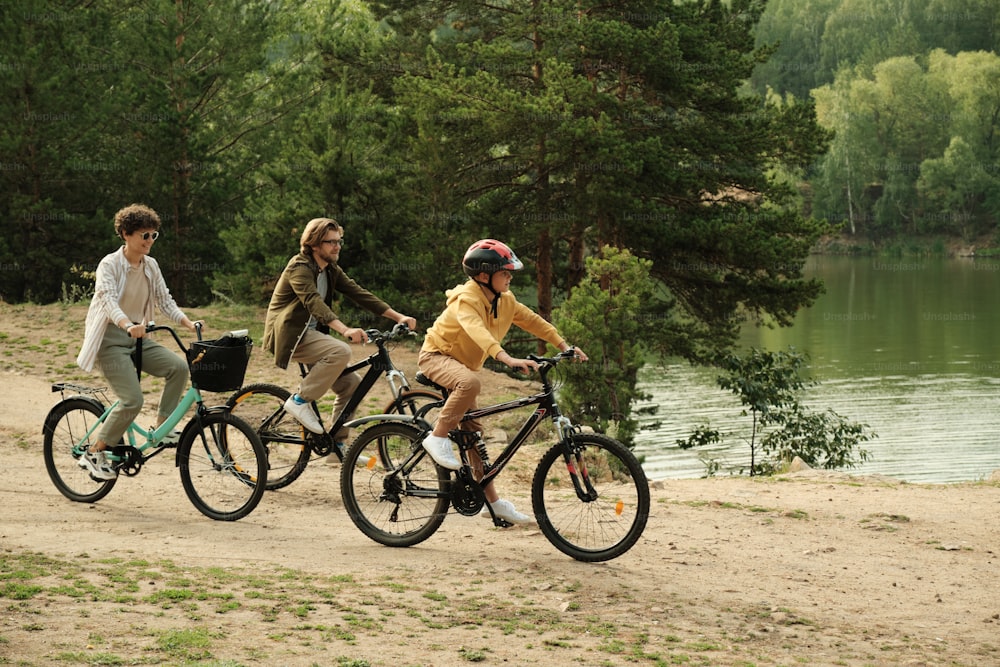 Contemporary happy family of three in casualawear sitting on bicycles while moving along riverside while enjoying active rest together