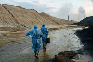 Rear view of two young contemporary female scientists in blue protective coveralls and rubber boots moving down dirty road and big puddles