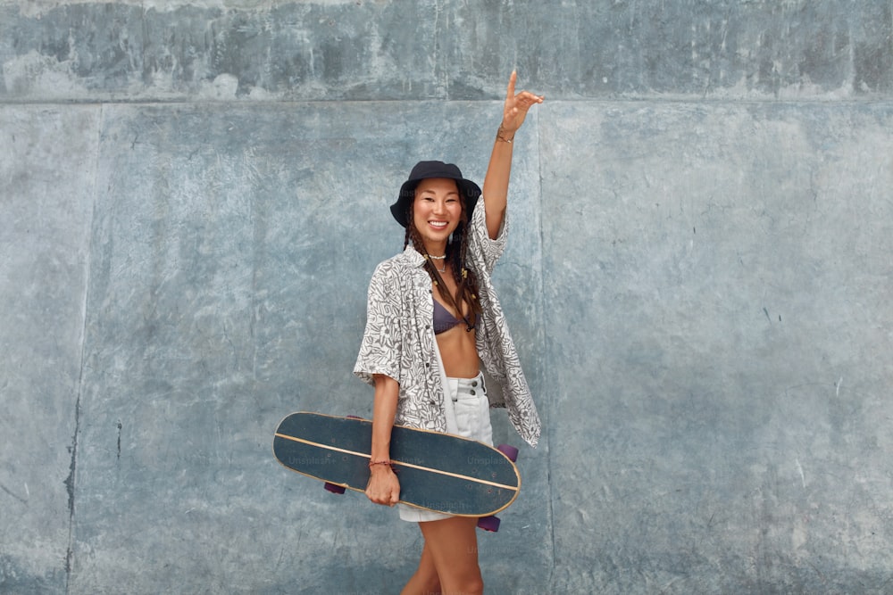 Skater Girl com retrato de skate. Adolescente asiático em traje casual posando contra parede de concreto no Skatepark. Subcultura urbana e skate como estilo de vida de adolescentes ativos na cidade.