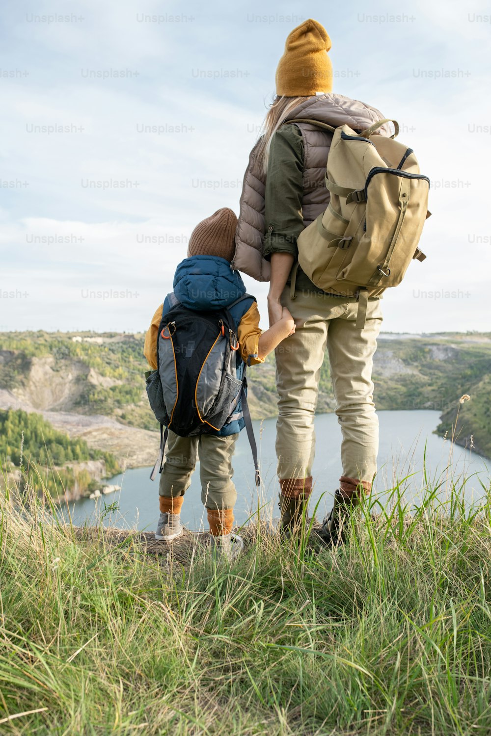 Rear view of young mother and little son with backpacks holding by hands while standing in front of lake surrounded by mountains during trip