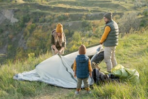 Young couple and their little son standing on green grass on top of mountain while putting tent on the ground in front of camera