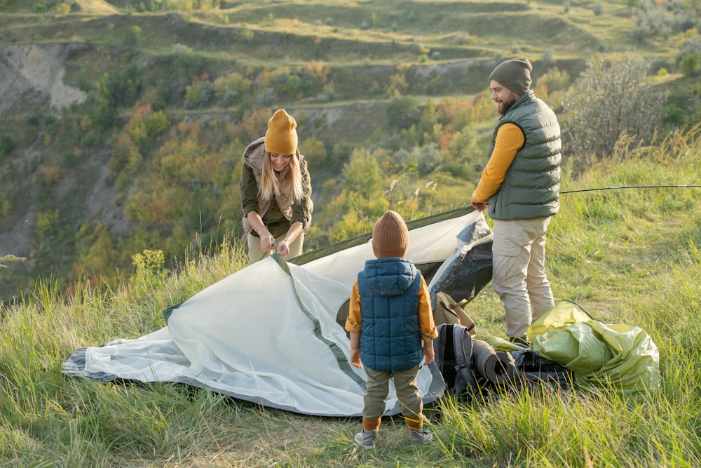 Young couple and their little son standing on green grass on top of mountain while putting tent on the ground in front of camera
