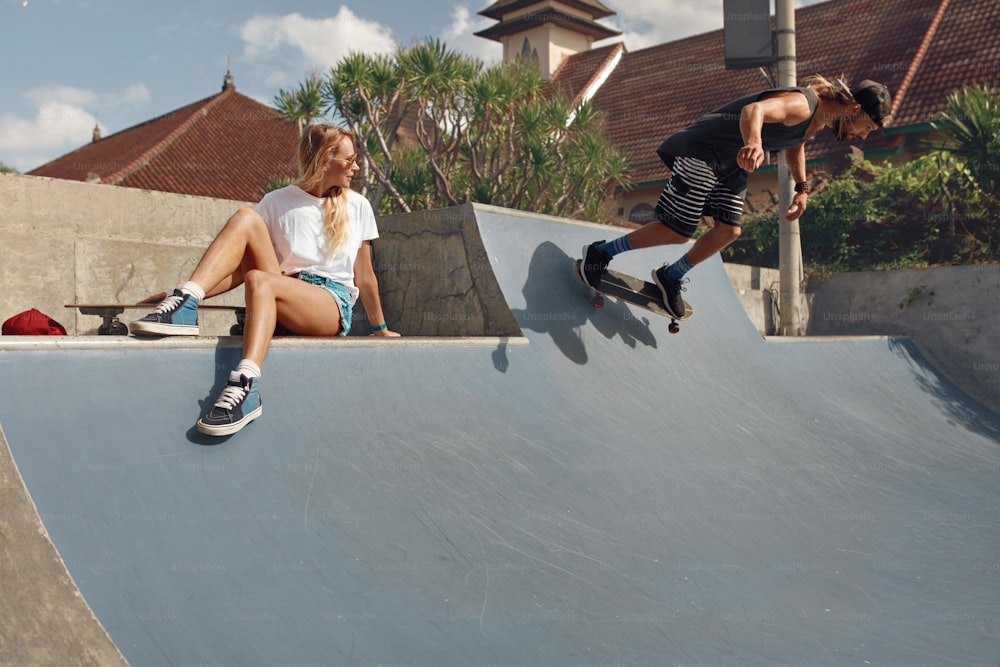 スケートパークのスケーターフレンズ。コンクリートのスロープに座っている女の子、スケートボードに乗っている男。街の夏のスケートボードのためのカジュアルな服装。都市生活のライフスタイルとしてのエクストリームスポーツ