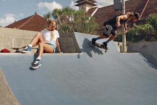 Skater Friends In Skatepark. Girl Sitting On Concrete Ramp, Guy Riding On Skateboard. Casual Outfit For Summer Skateboarding In City. Extreme Sport As Lifestyle Of Urban People.