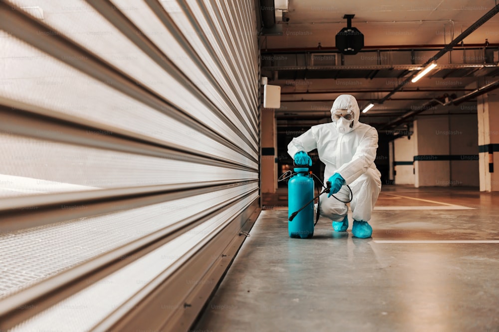 Man in protective sterile uniform crouching and disinfecting garage with disinfectant.