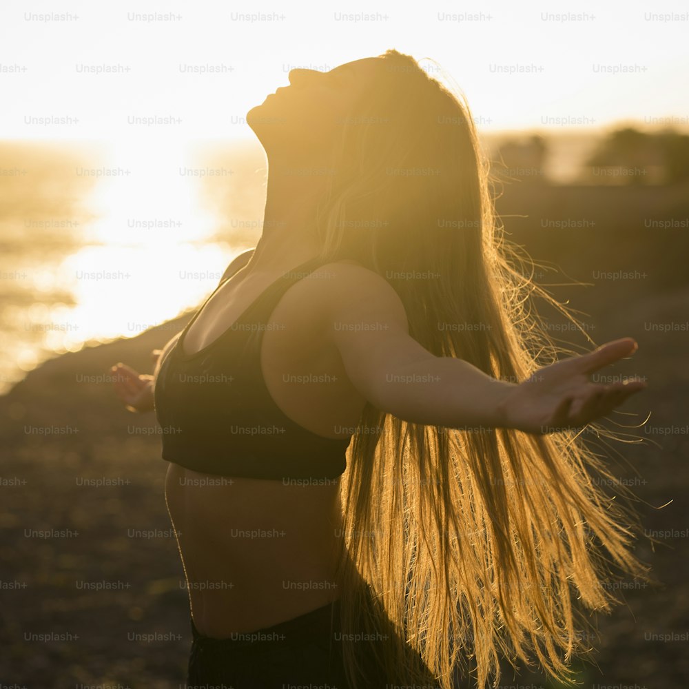 若いアスリートの健康的なフィットネスの女性は、夕日を腕を広げて呼吸を楽しんでいます - フィットネスライフスタイルの女の子と海と海岸の景色の黄金の日光 - スポーツ野外活動のコンセプト