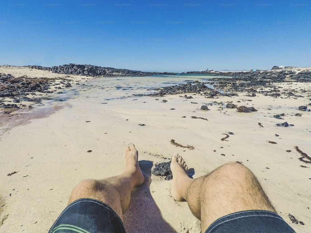Punto di vista alternativo delle gambe dell'uomo in spiaggia con il costume da bagno che si gode la sabbia e l'acqua blu e il cielo durante le vacanze estive