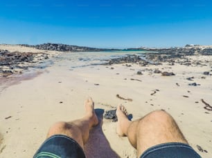 Ponto de vista alternativo das pernas do homem na praia com roupa de banho apreciando a areia e a água azul e o céu nas férias de verão