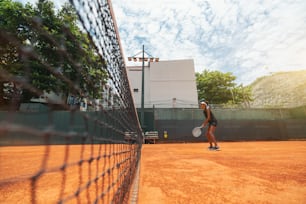 Wide-angle view of a young slim biracial female with a racket in hands standing on a clay court surface and having a tennis play round or a warm-up workout; a net on the left, warm sunny day, Brazil