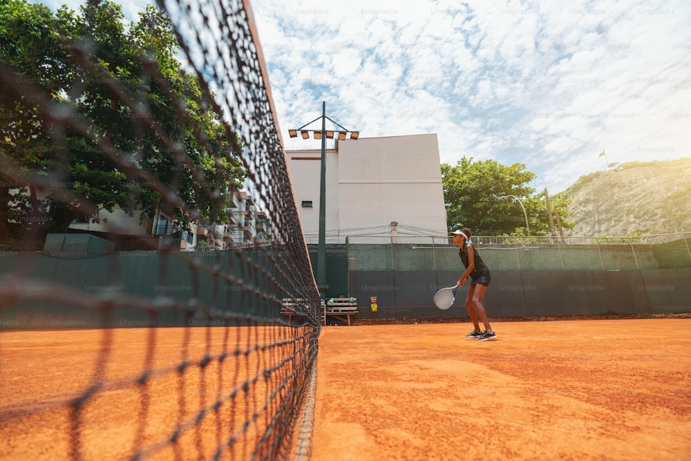 Wide-angle view of a young slim biracial female with a racket in hands standing on a clay court surface and having a tennis play round or a warm-up workout; a net on the left, warm sunny day, Brazil
