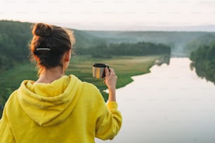 Young woman girl traveler in yellow hoodie with cup of the morning coffee looking at sunrise on beautiful view. Local tourism, weekend trip