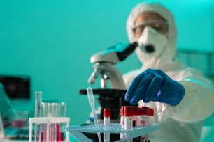 Gloved hand of researcher in protective coveralls and respirator sitting by microscope while taking one of small flasks with blood sample