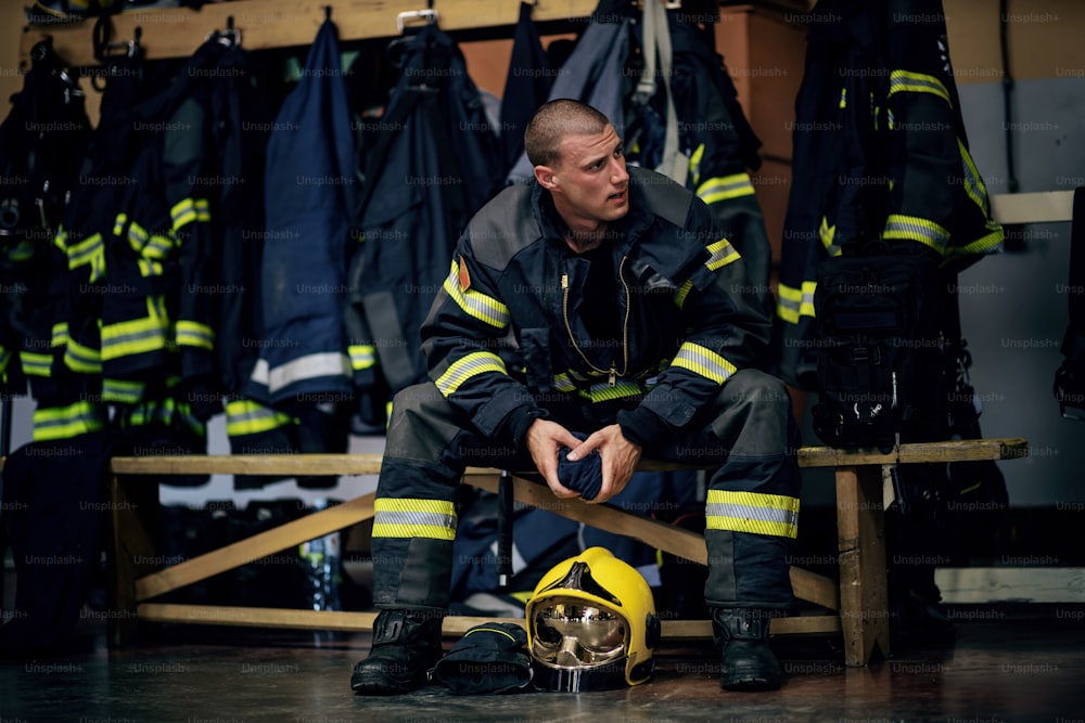 Junger attraktiver Feuerwehrmann in Schutzuniform sitzt in der Feuerwache und wartet auf andere Feuerwehrleute. Er ist bereit zum Handeln.
