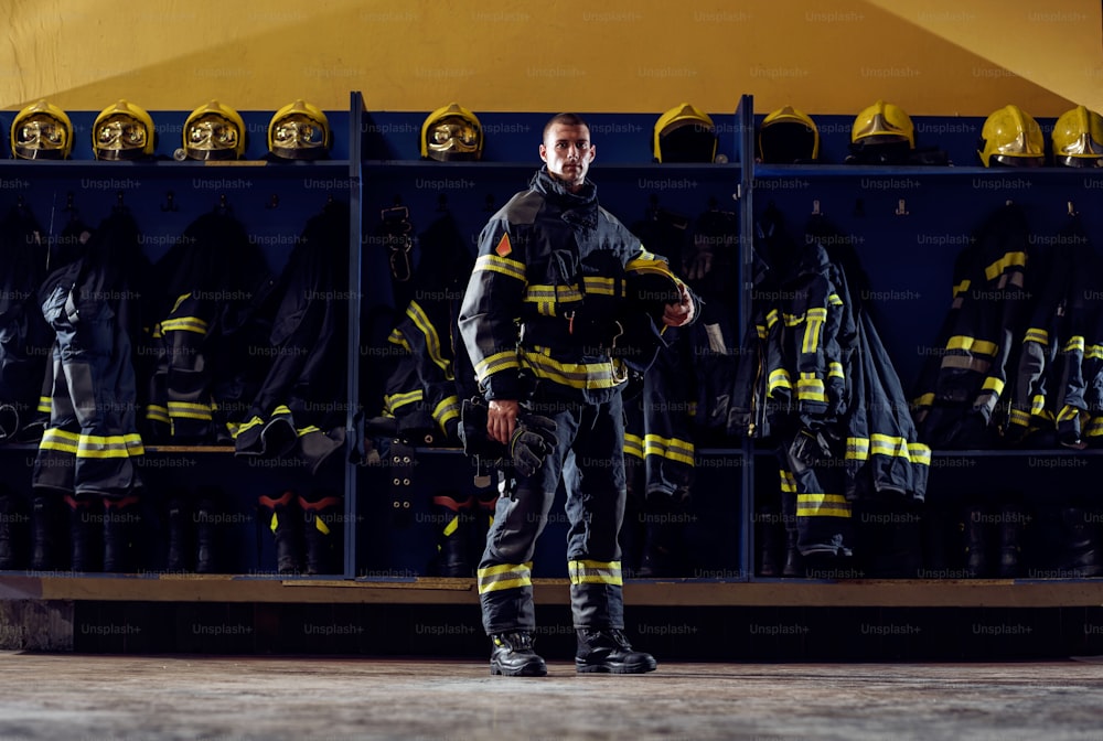 Brave young firefighter standing in fire station in protective uniform and holding helmet under armpit.