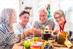 Cheerful family with mixed generations enjoy together the Christmas winter eve lunch at home with chocolate and cakes and frees - people celebrate event in friendship and love - white home indoor and wooden table