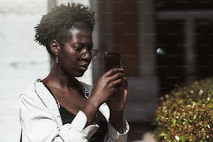 Portrait of a young beautiful black woman in a white trench and spectacles taking pics outdoors using the camera of her smartphone; a charming African girl photographing attractions via the cellphone