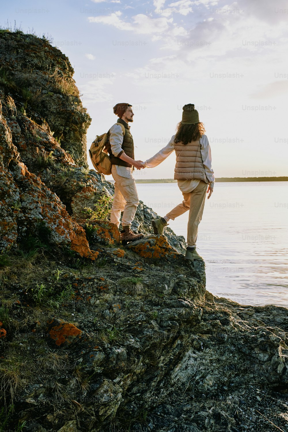 Liebevolle Wanderer in Freizeitkleidung, die auf Felsen der Klippe stehen und auf den See schauen, während sie bei Sonnenuntergang zusammen spazieren gehen