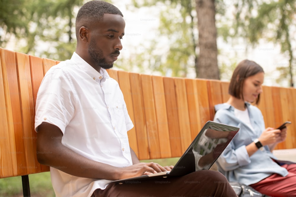 Serious guy of African ethnicity in casualwear sitting on bench in park and networking while looking at laptop display on background of girl
