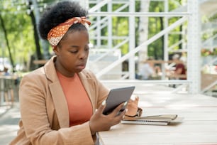 Young elegant businesswoman of African ethnicity scrolling in mobile gadget while sitting by table in cafe and planning working day
