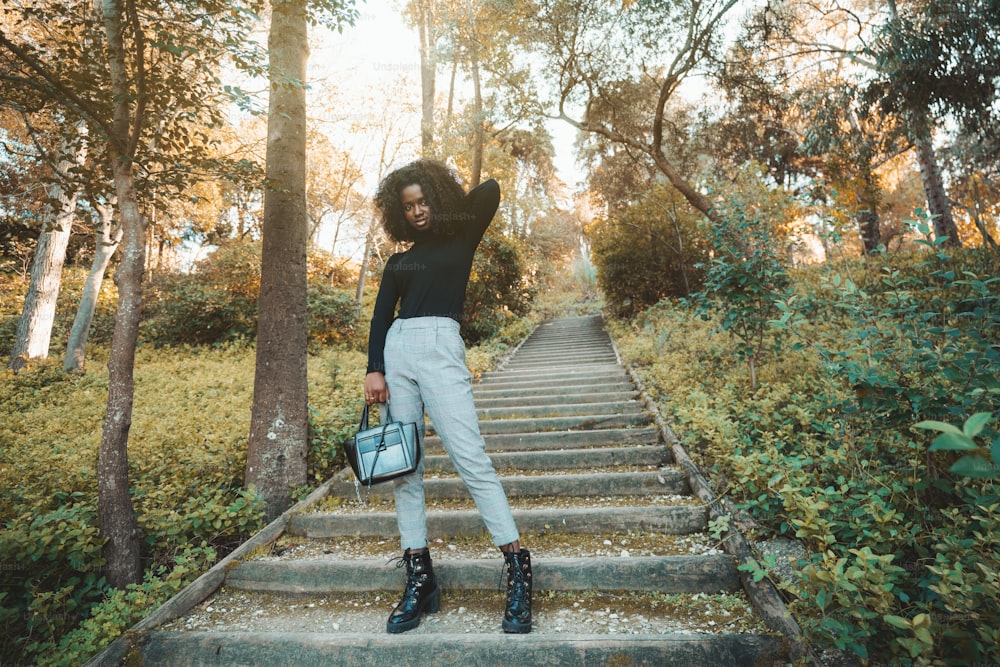 A young charming African female in a black turtleneck and plaid trousers, with a small bag in hand, is staying on old wooden stairs in an autumn park; a copy space place on the right; wide-angle shot