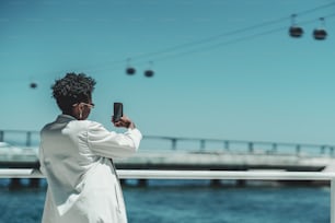 Vista desde atrás de una elegante joven negra con cabello afro rizado con elegantes gafas de sol y una gabardina blanca está de pie al aire libre y tomando una selfie o fotografiando con el teléfono celular un teleférico