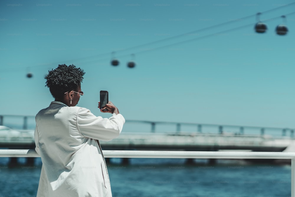 View from behind of an elegant young black female with curly afro hair in stylish sunglasses and a white trench is standing outdoors and taking a selfie or photographing on the cellphone a ropeway