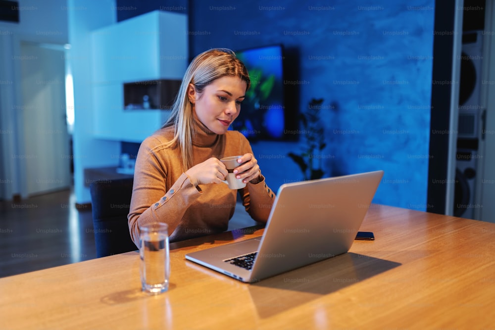 Young dedicated stunning blond female entrepreneur sitting at home, drinking coffee and working on important project during covid 19 outbreak.