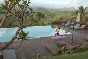 Beautiful Girl Doing Yoga On Poolside In Morning In Bali, Indonesia. Young Woman In Sportswear Standing In Warrior Pose Or Virabhadrasana On Sport Mat Near Infinity Pool Against Tropical Landscape.