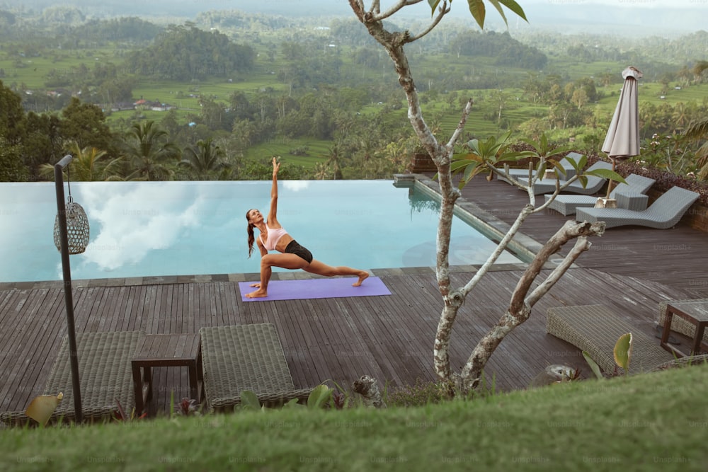 Beautiful Girl Doing Yoga On Poolside In Morning In Bali, Indonesia. Young Slim Woman In Sportswear Standing In Side Angle Pose On Sport Mat Near Infinity Pool Against Tropical Landscape.