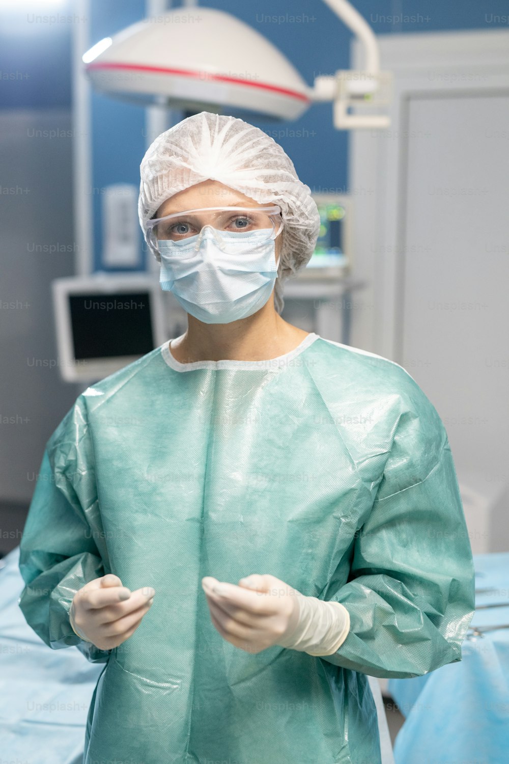 보호 장갑, 마스크, 안경, 작업복을 입은 젊은 현대 여성 외과의가 수술실에서 카메라 앞에 서 있다